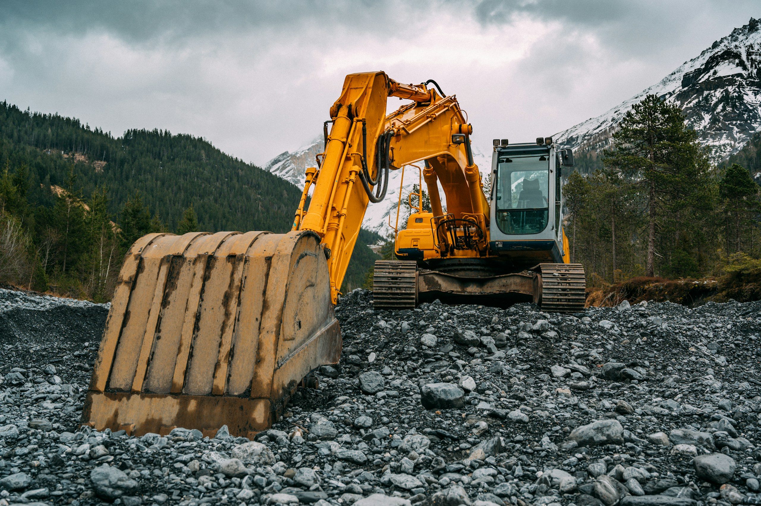 Yellow excavator on a rocky site below snow-covered mountains